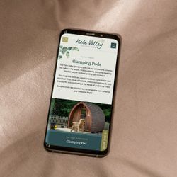 Smartphone displaying Hele Valley Holiday Park website showing glamping pod information, placed on a beige fabric surface.