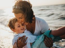 Mother smiling and holding her young son while standing on a beach at sunset.