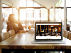 Laptop on wooden table displaying a restaurant website in a bright, sunlit cafe.