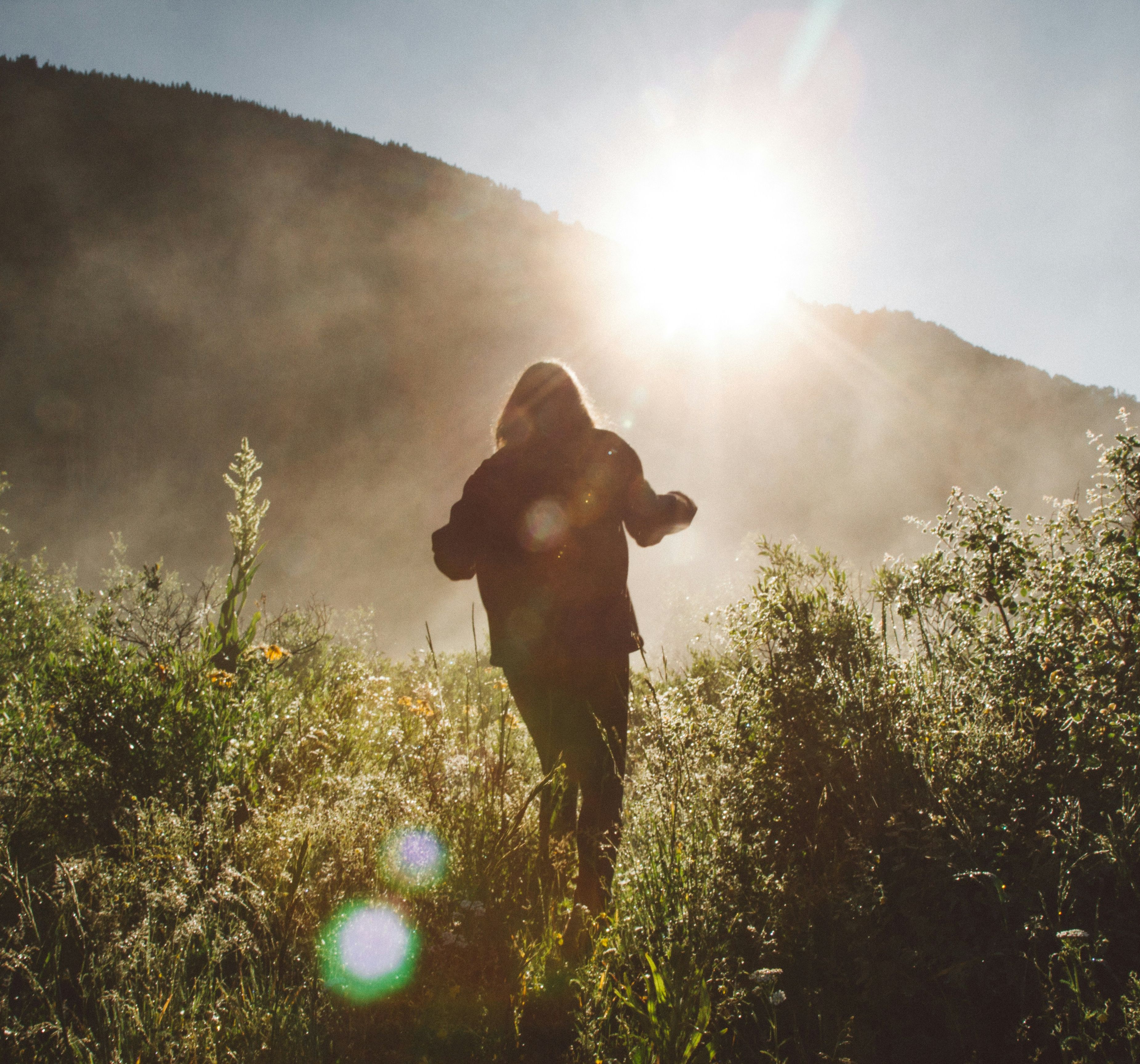 Person walking through tall grass in a sunlit field surrounded by mountains