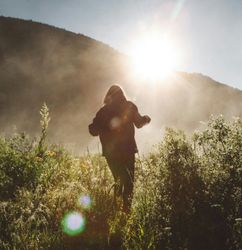 Person walking through tall grass in a sunlit field surrounded by mountains