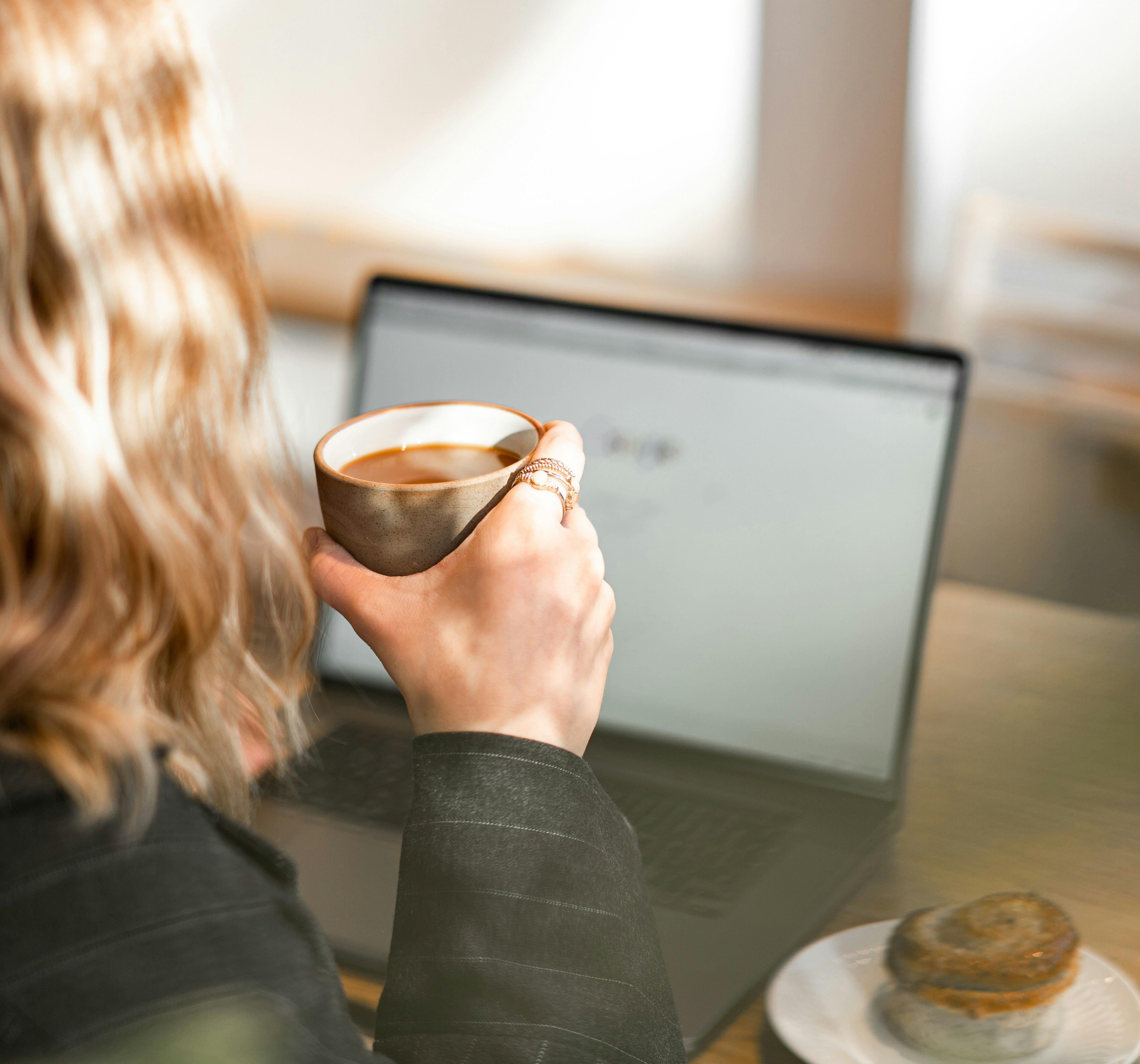 Person holding a cup of coffee in front of a laptop with a pastry on a plate nearby