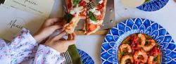 Top view of people sharing pizza with fresh basil and a plate of shrimp with tomato sauce on a blue patterned plate