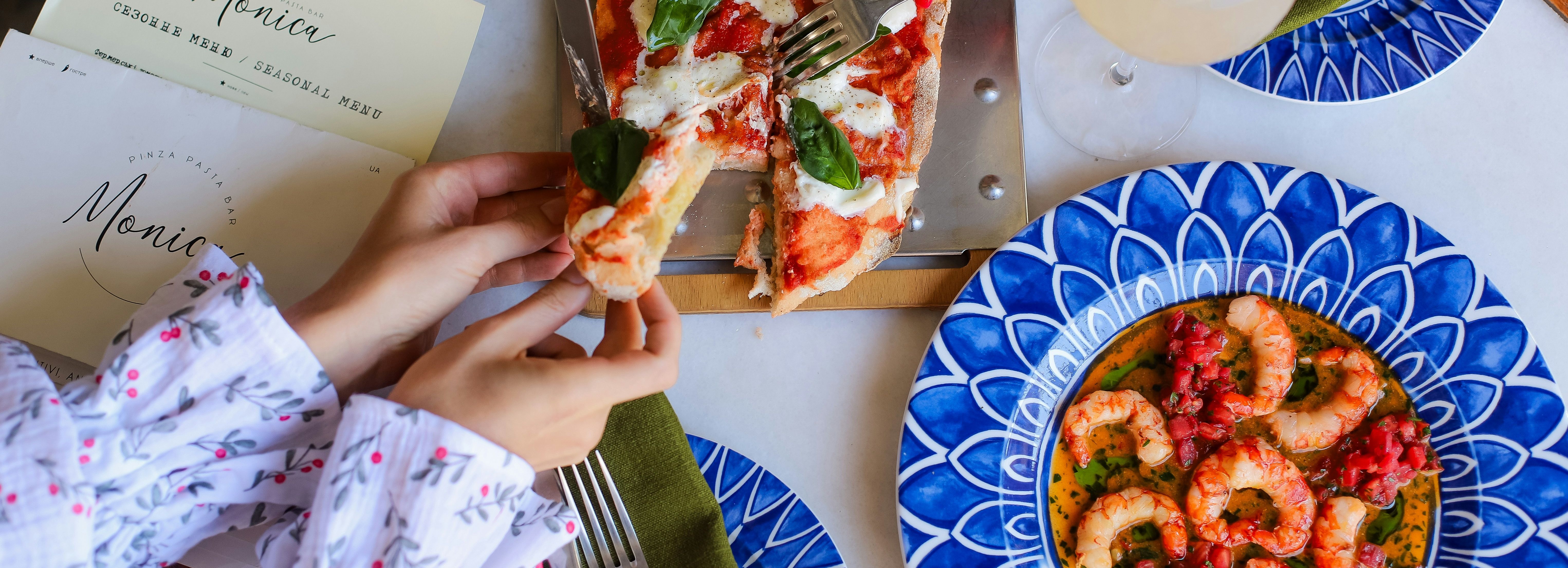 Top view of people sharing pizza with fresh basil and a plate of shrimp with tomato sauce on a blue patterned plate