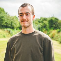 Young man standing outside on a grassy field with trees in the background