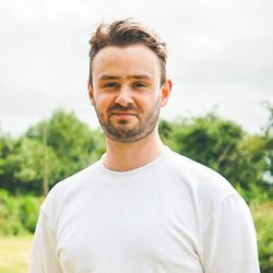 Man with short brown hair and beard wearing a white t-shirt standing outdoors