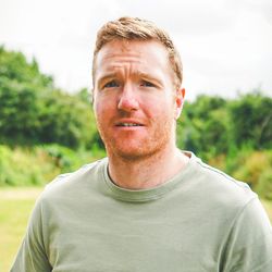Man with reddish hair and tattoos on his arms standing outdoors in front of greenery