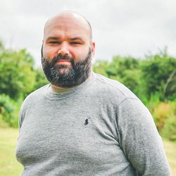 Man with a beard and shaved head standing outside in a grey sweater with greenery in the background