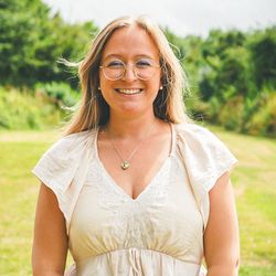 Smiling woman with long blonde hair, wearing glasses and a cream top, standing outdoors in a grassy field.