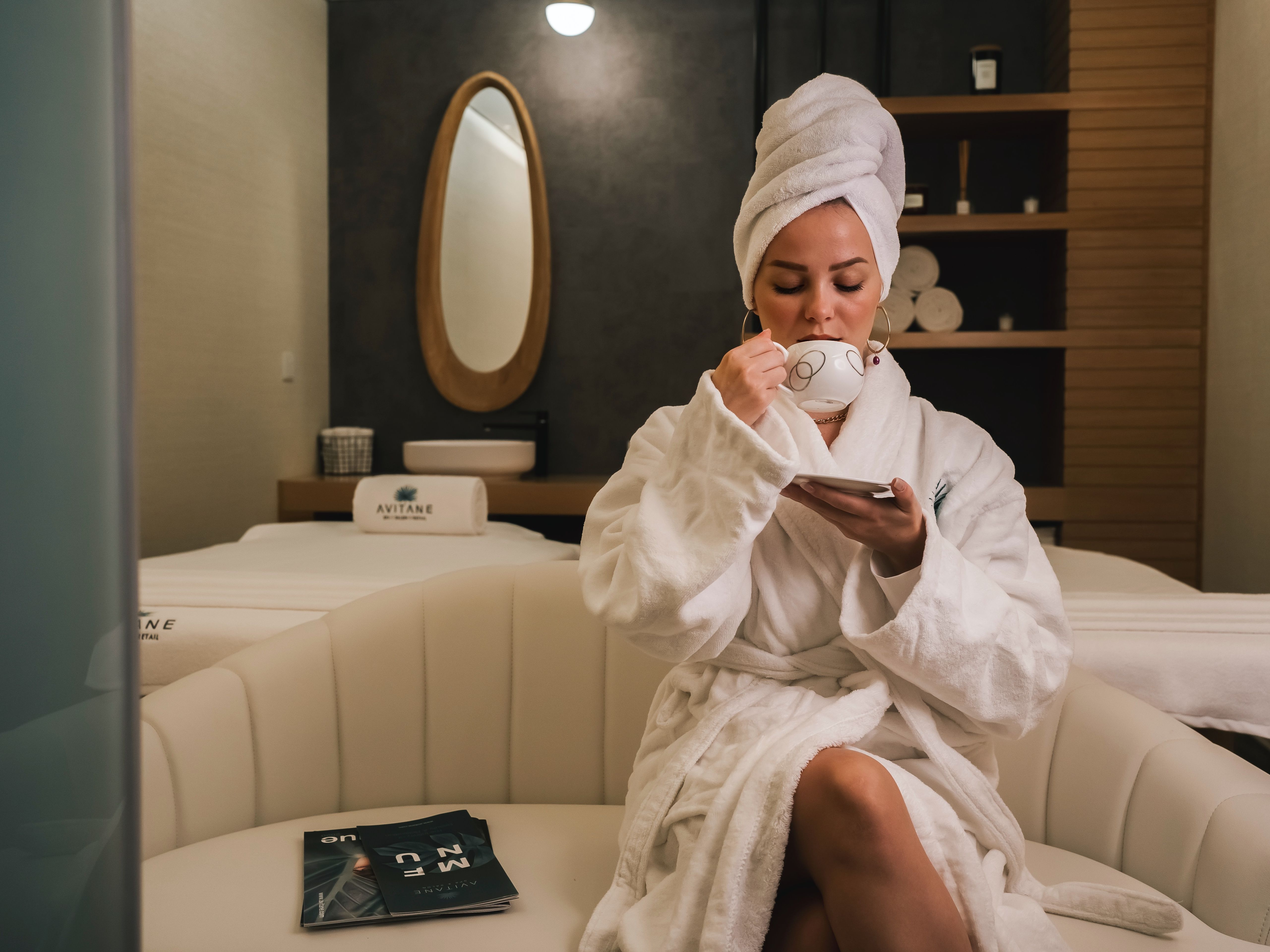 Woman relaxing in a spa lounge wearing a white robe and towel on her head, sipping tea.