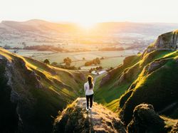 Person standing on grassy cliff overlooking a sunlit valley and rolling hills at sunrise or sunset.