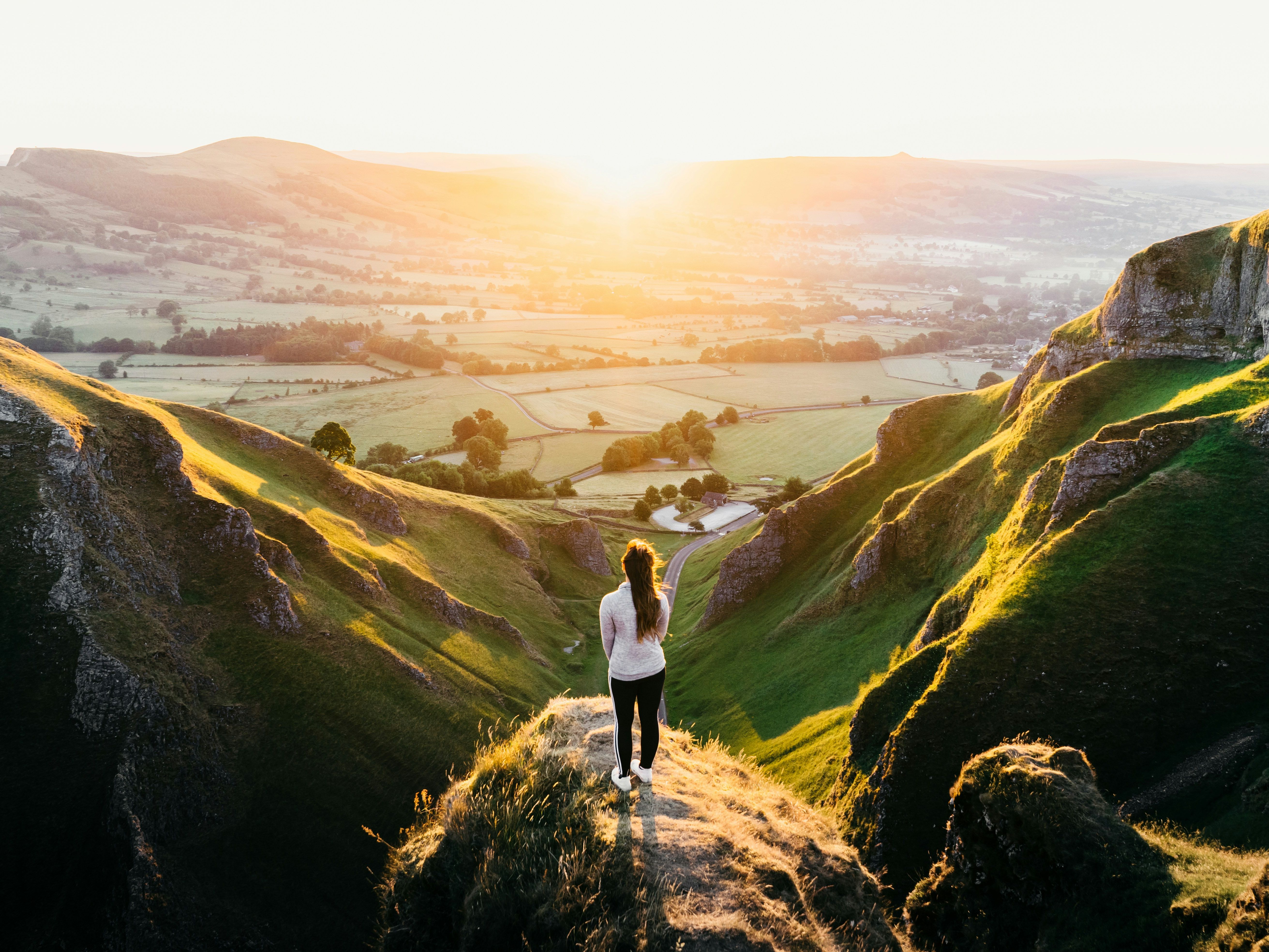 Person standing on grassy cliff overlooking a sunlit valley and rolling hills at sunrise or sunset.