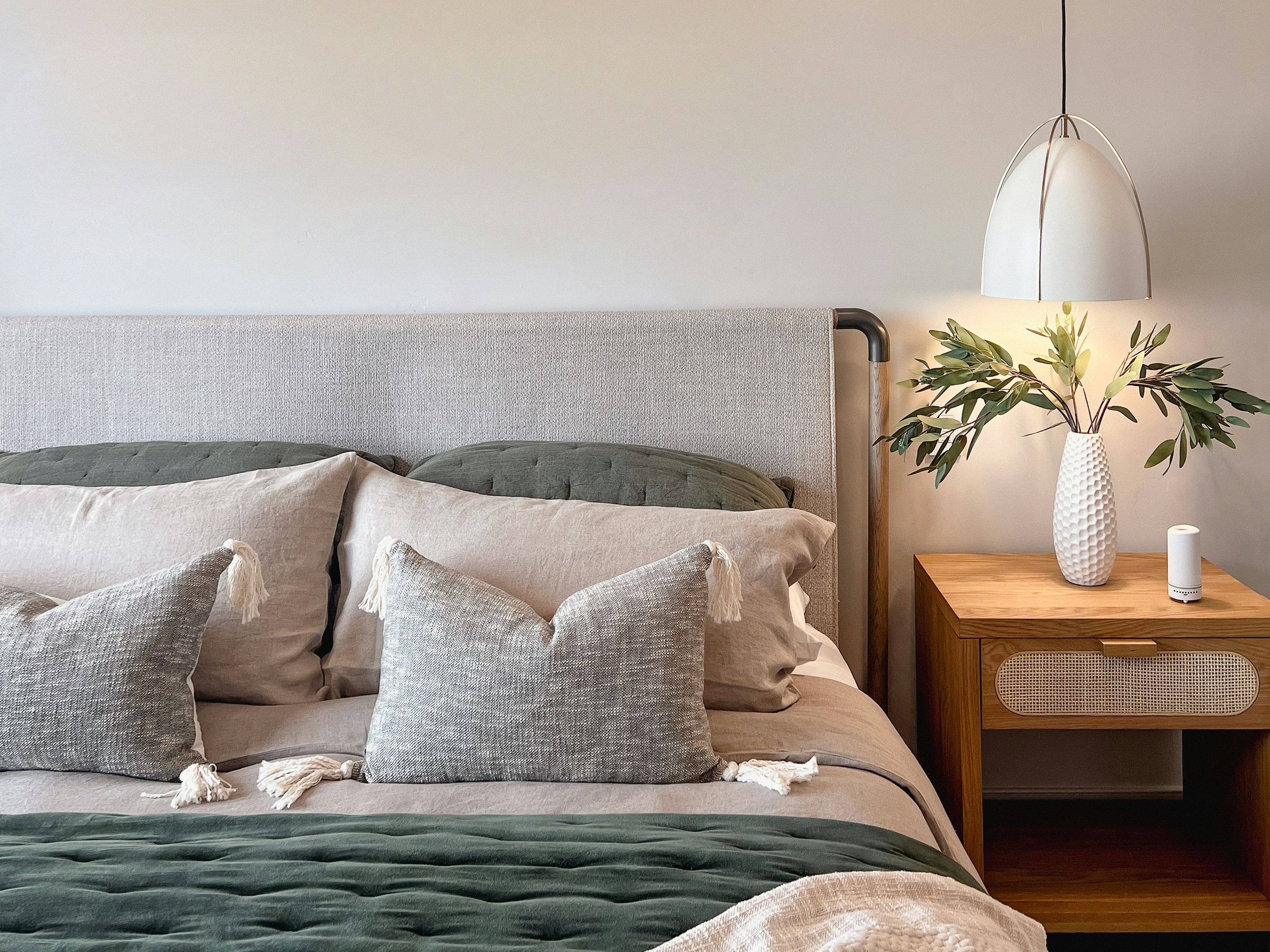 Modern bedroom with neutral tones, green accents, and a bedside table with a lamp and vase of leaves