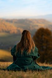Person with long hair wearing a green coat sitting on grass and looking at a scenic mountain landscape