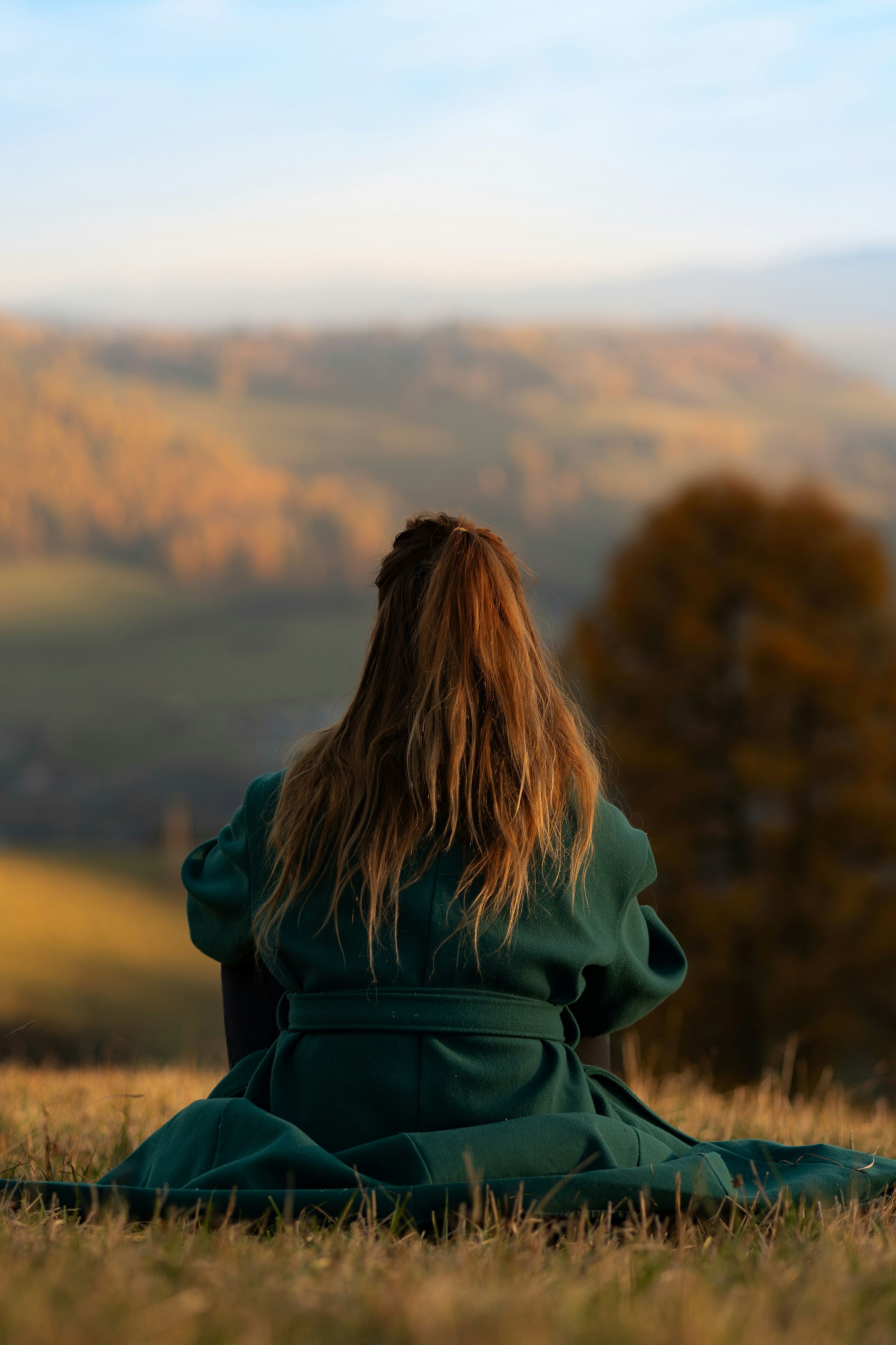 Person with long hair wearing a green coat sitting on grass and looking at a scenic mountain landscape