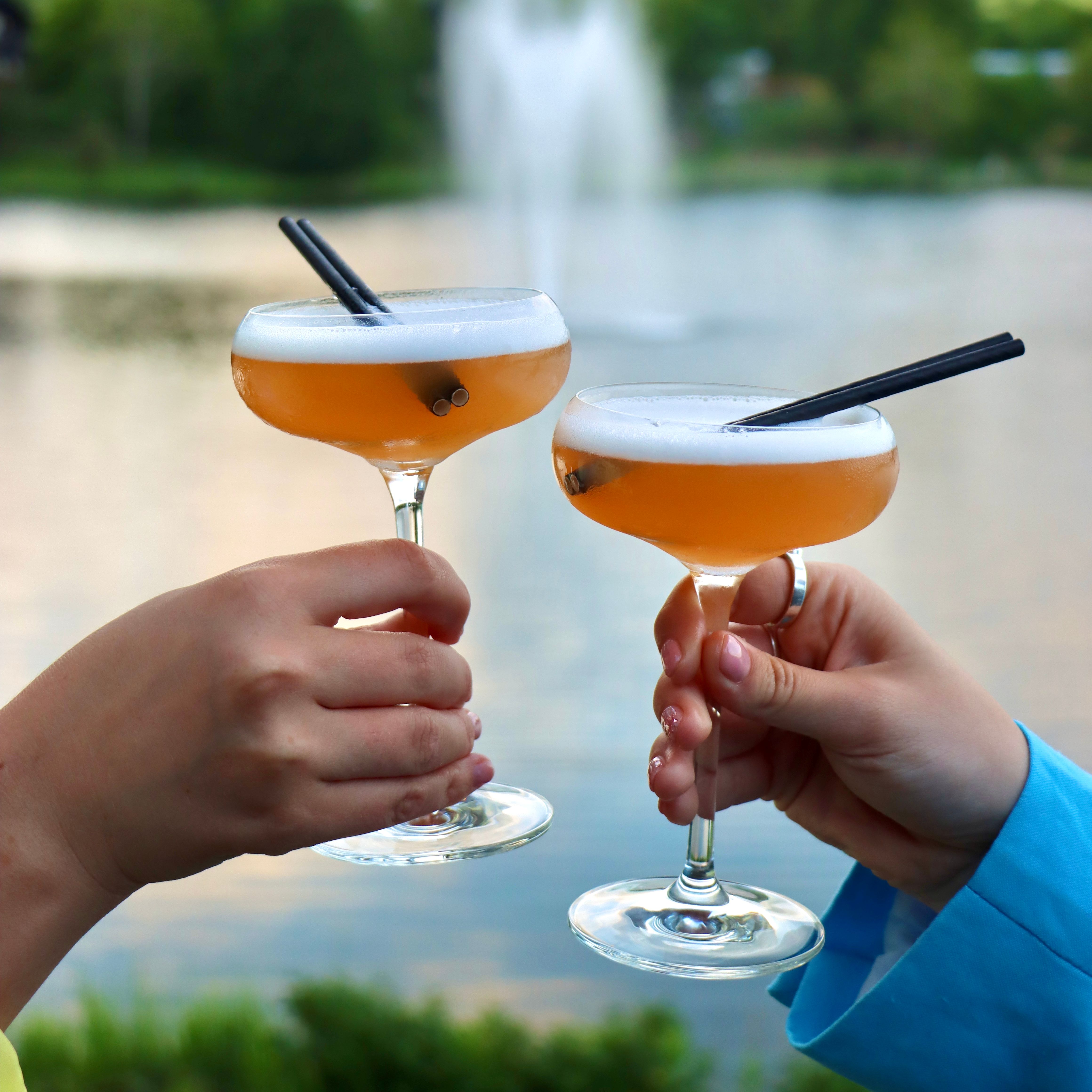 Two people toasting with cocktail glasses near a lake with a fountain in the background.