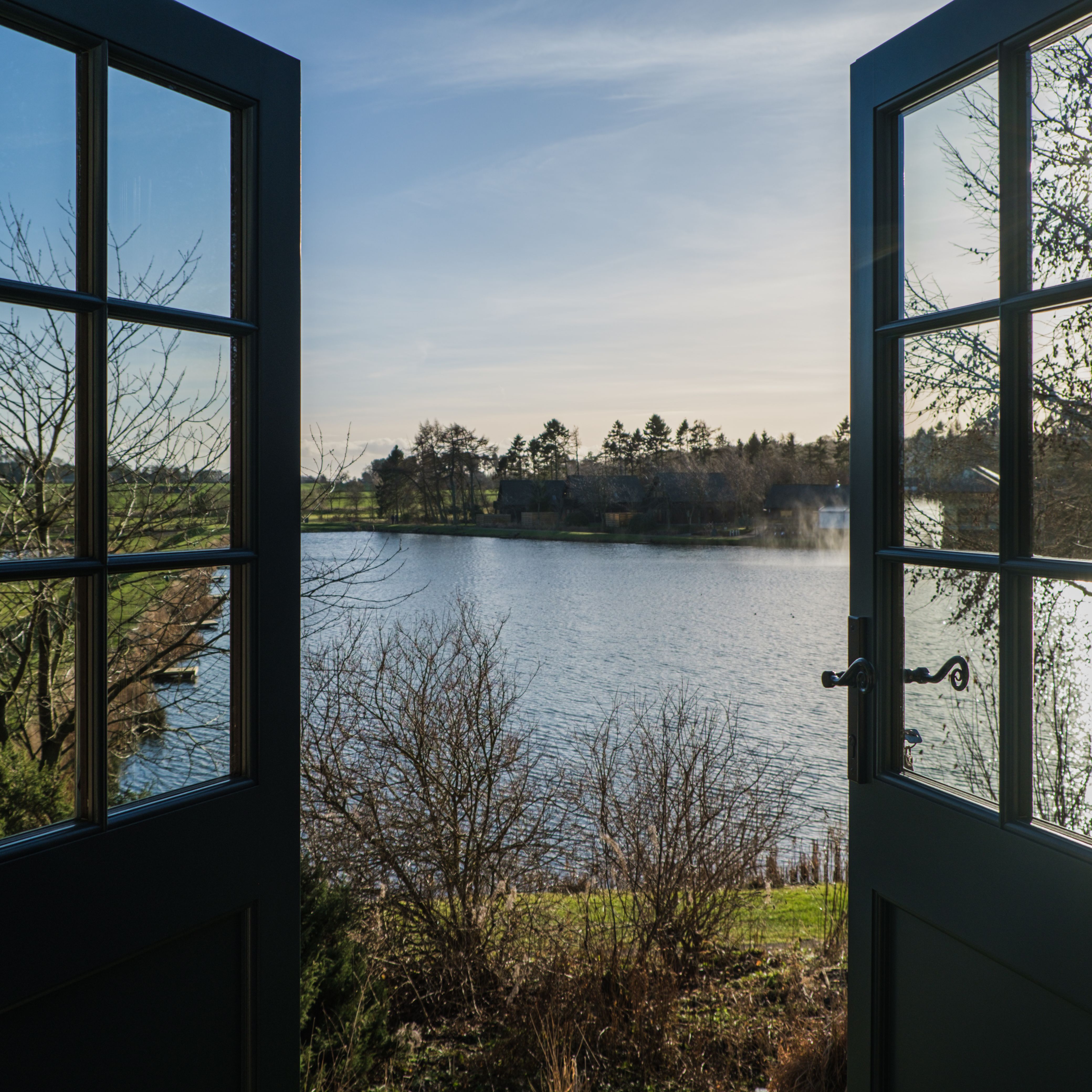 Open french doors with a view of a peaceful lake and trees under a clear sky.