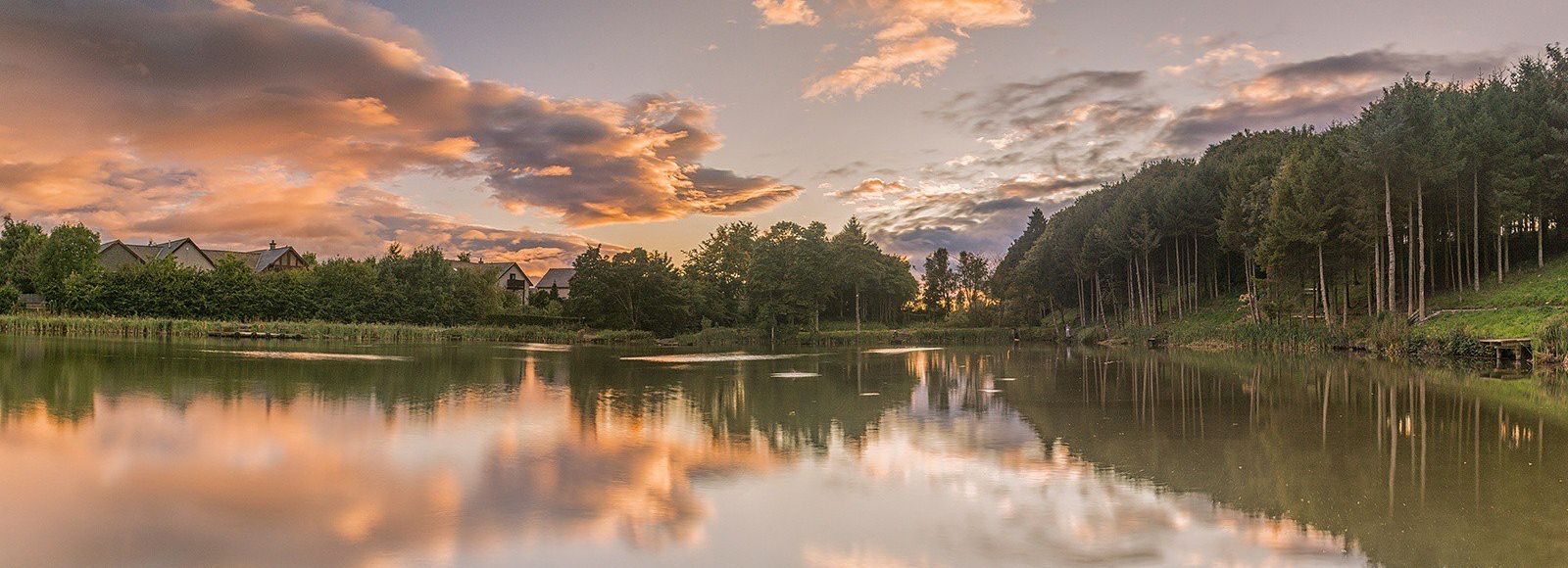 Serene lake reflecting trees and colorful clouds during sunset with houses in the background.