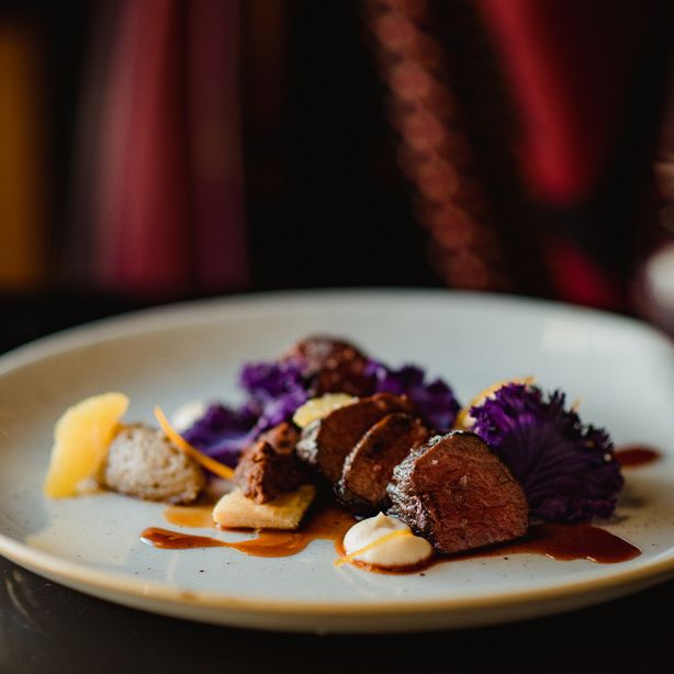 Gourmet plated dish with sliced meat, sauces, and purple garnish on a white plate