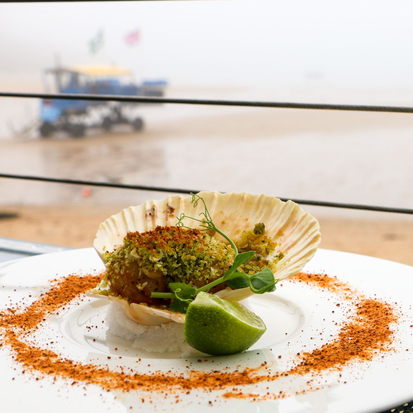 Gourmet baked scallop dish served in a shell with lime wedge on a white plate, garnished with herbs and spices, set against a beach background.
