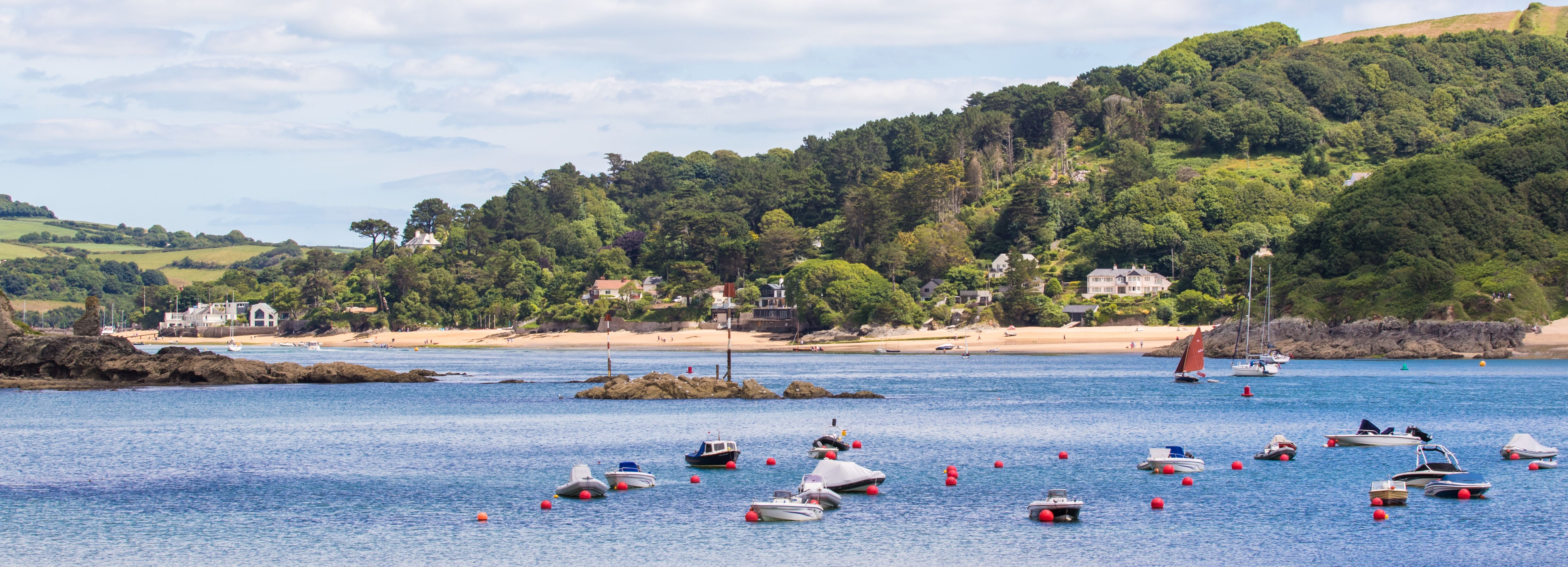 Small boats floating on calm water near a sandy beach with green hills and houses in the background