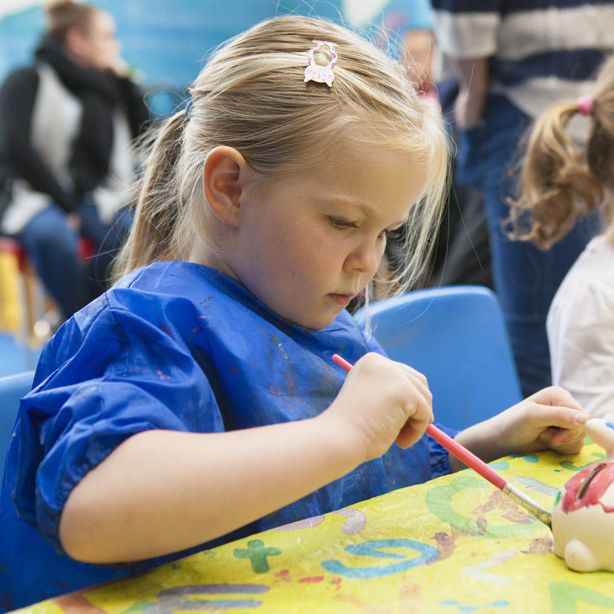 Young girl painting a ceramic figure at a table with another child beside her.