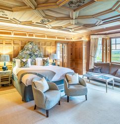 Luxury bedroom with wood paneling, ornate ceiling, large bed, armchairs, and a sitting area with a sofa near a window.