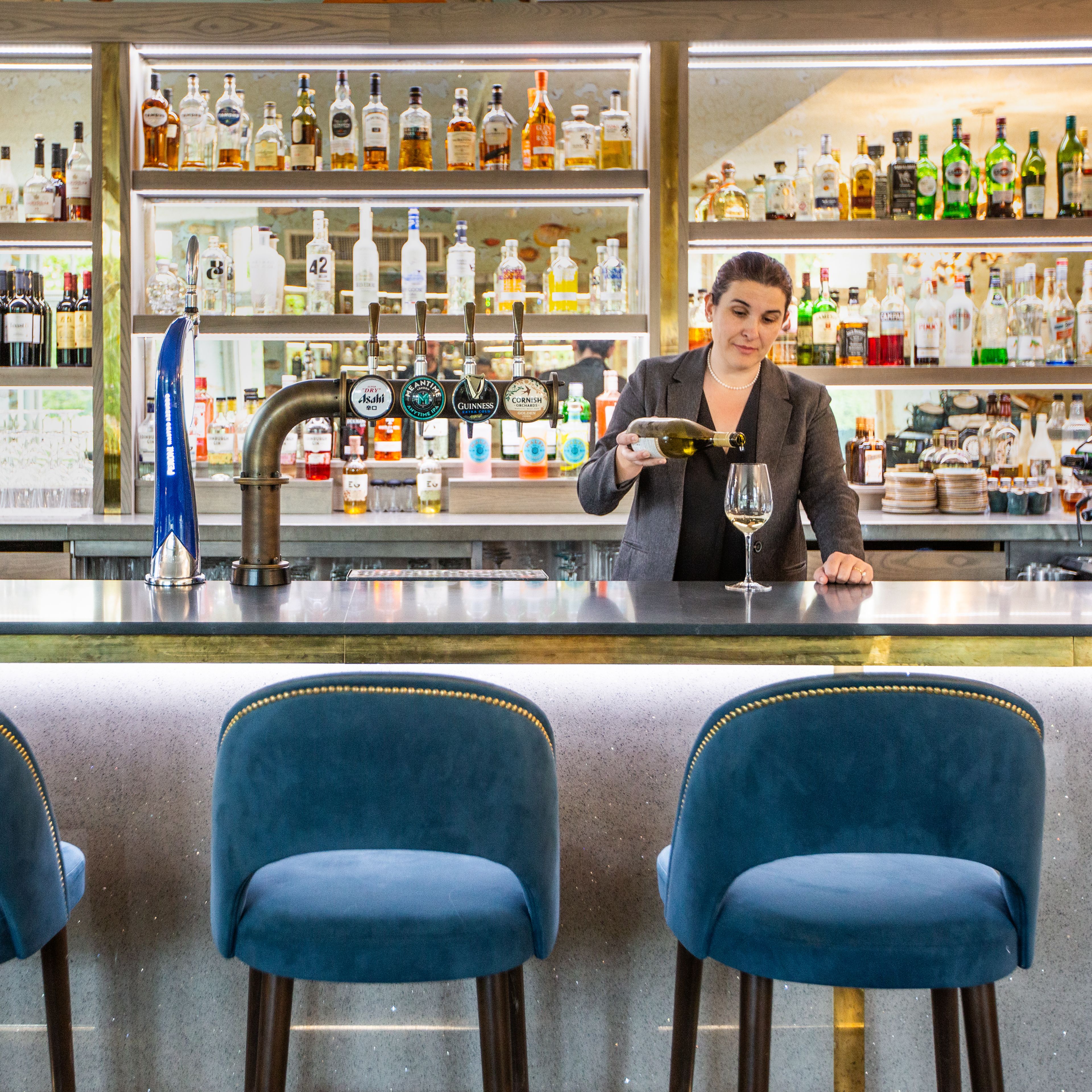 Bartender pouring wine behind a modern bar with blue stools and shelves of liquor bottles