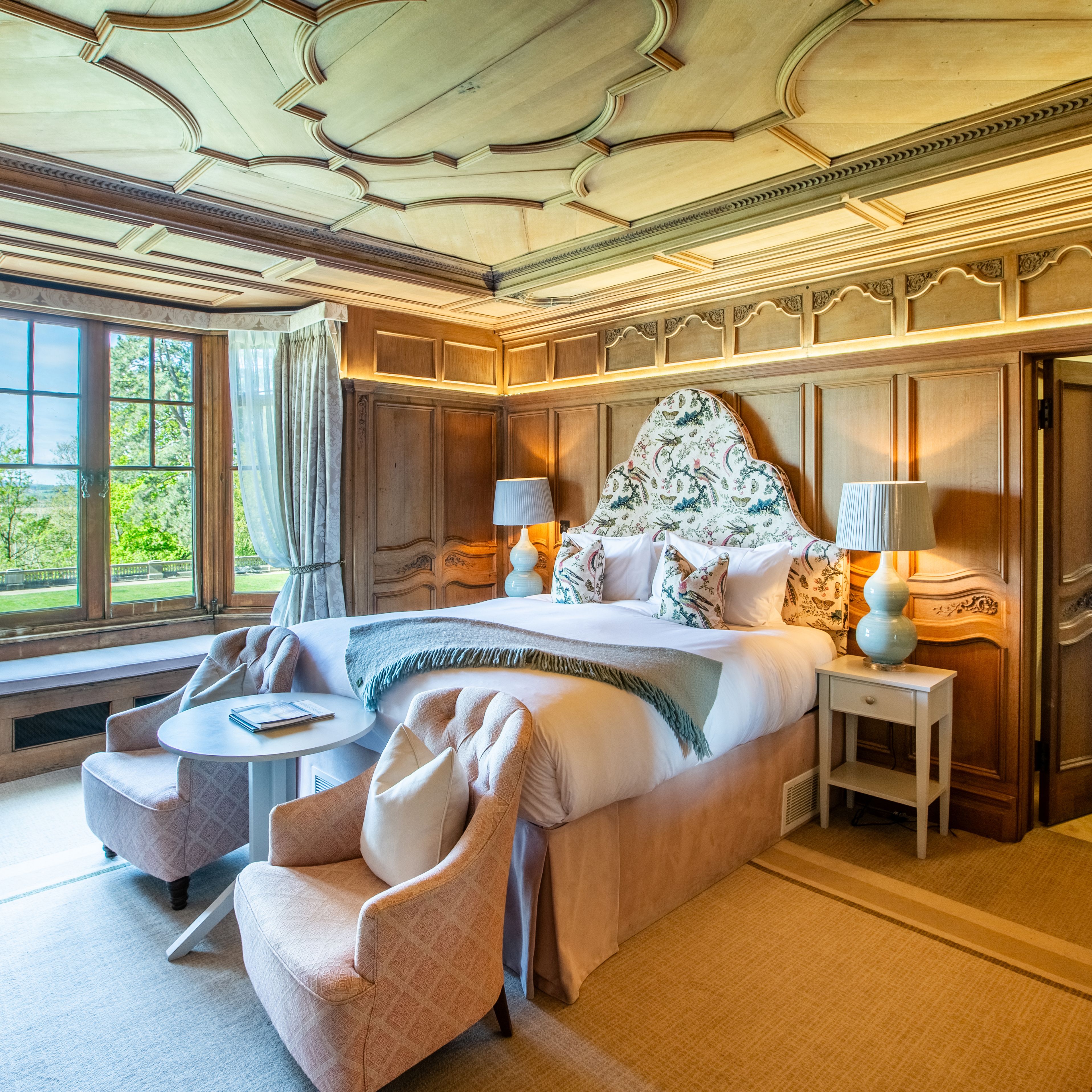 Elegant bedroom with wooden paneling, large bed, decorative headboard, and sitting area by a large window.