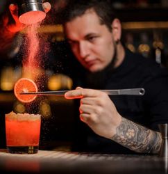 Bartender garnishing a red cocktail with powdered garnish and a dried citrus wheel