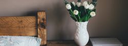 White tulips in a decorative vase on a rustic wooden nightstand next to a bed.