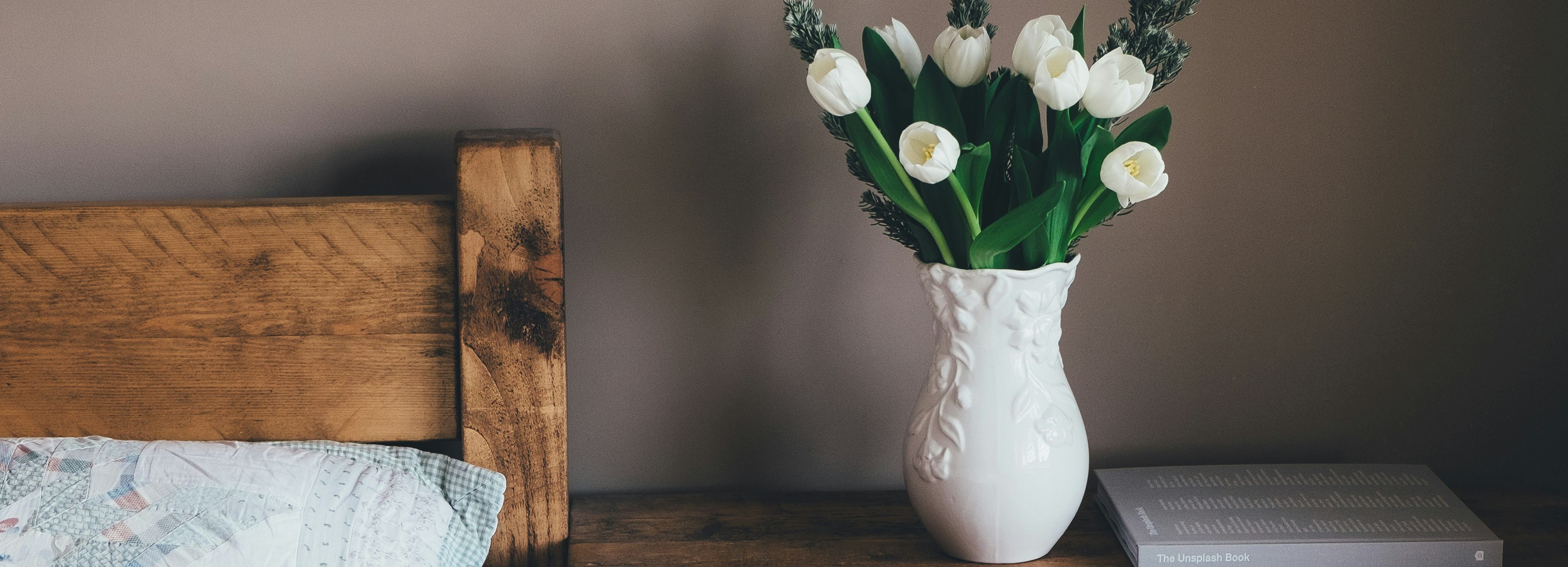 White tulips in a decorative vase on a rustic wooden nightstand next to a bed.