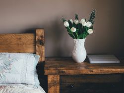 White tulips in a decorative vase on a rustic wooden nightstand next to a bed.