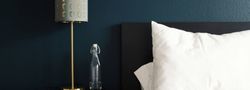 Modern bedroom setting with a gold lamp, glass water bottle, white pillow, and dark blue wall.