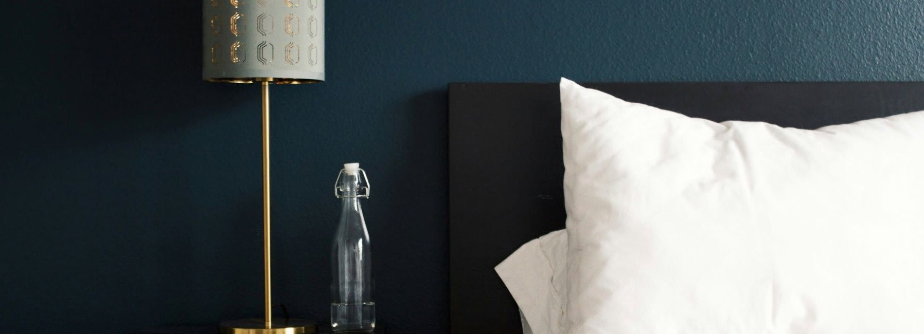 Modern bedroom setting with a gold lamp, glass water bottle, white pillow, and dark blue wall.