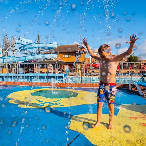 Child playing in water jets at an outdoor splash park with a blue water slide in the background.