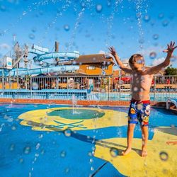 Child playing in water jets at an outdoor splash park with a blue water slide in the background.