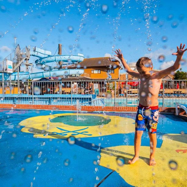 Child playing in water jets at an outdoor splash park with a blue water slide in the background.