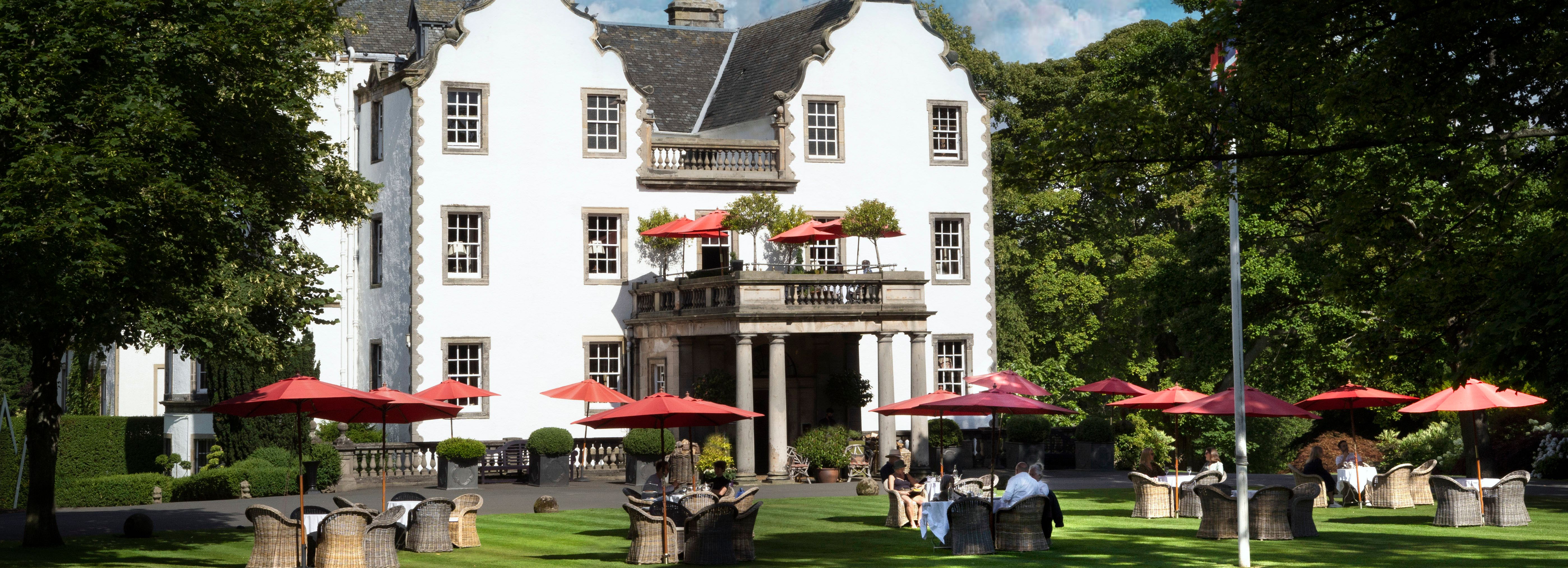 Elegant white manor house surrounded by lush gardens with outdoor seating and red umbrellas.