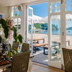 Indoor dining area with open glass doors leading to an outdoor terrace overlooking the water and a scenic coastline.