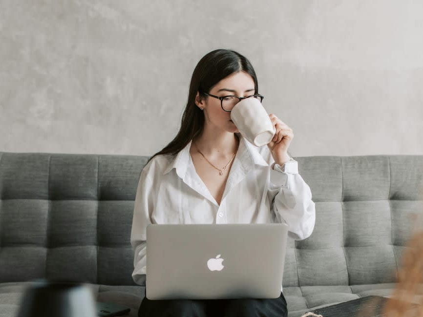 Woman sitting on a couch using a laptop and drinking from a mug