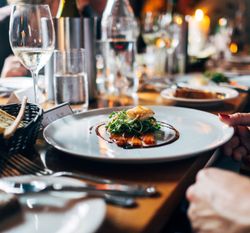 A beautifully plated gourmet dish on a white plate at a restaurant table, surrounded by glasses of wine and bread.