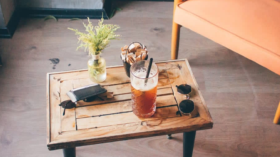 Wooden table with iced drink, sunglasses, wallet, and a small vase next to an orange chair