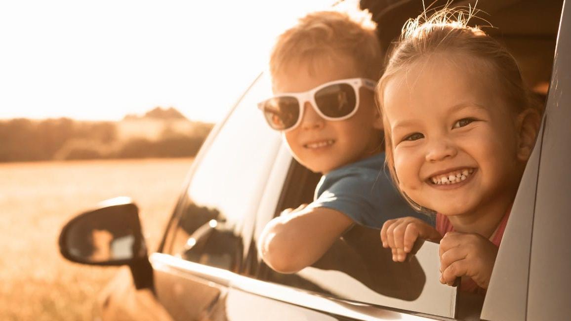 Two smiling children leaning out of a car window on a sunny day