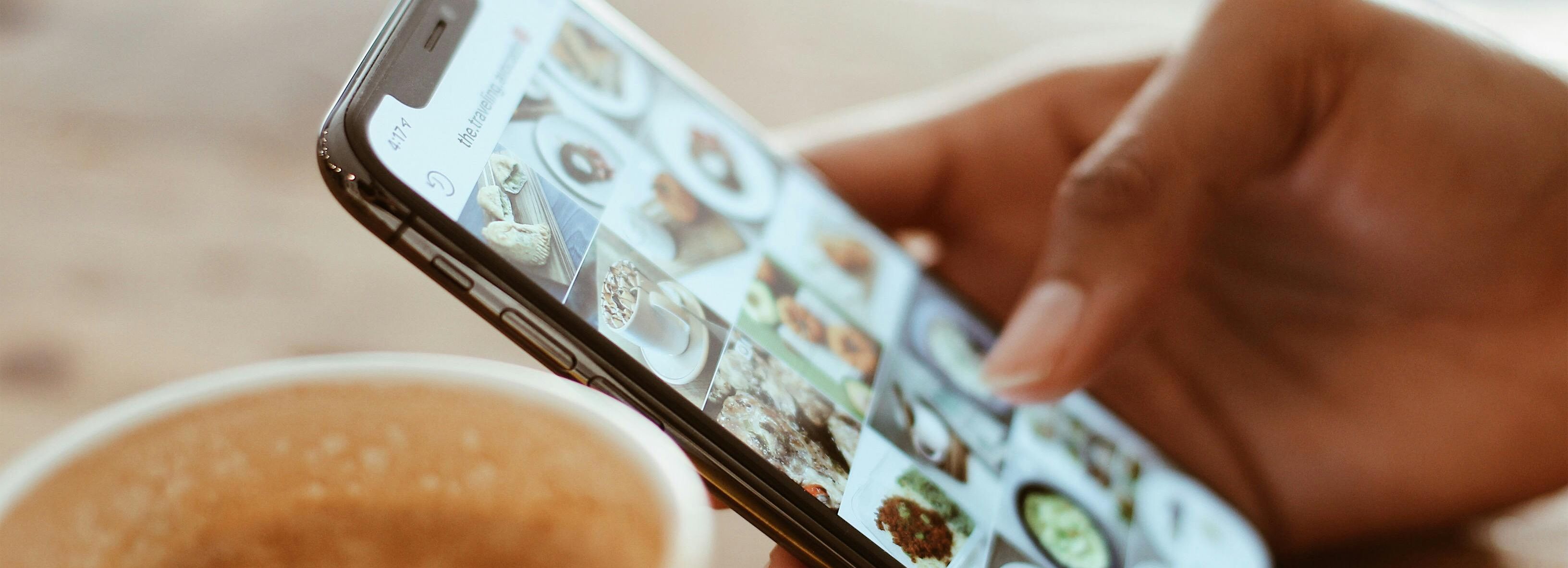 Person browsing food photos on a smartphone next to a cup of coffee