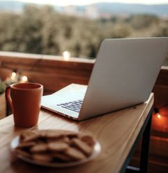 Laptop, coffee mug, and plate of cookies on a wooden table on a cozy balcony with string lights.