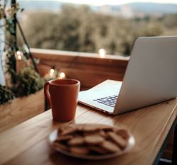 Laptop, coffee mug, and plate of cookies on a wooden table on a cozy balcony with string lights.