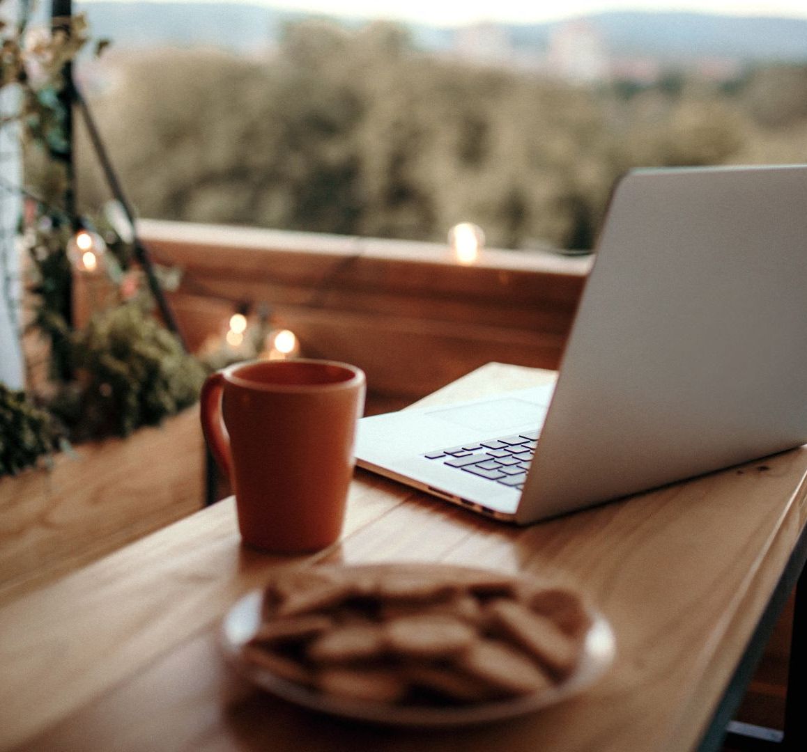 Laptop, coffee mug, and plate of cookies on a wooden table on a cozy balcony with string lights.