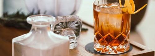 A glass of cocktail with ice and an orange peel garnish on a wooden tray next to a decanter.
