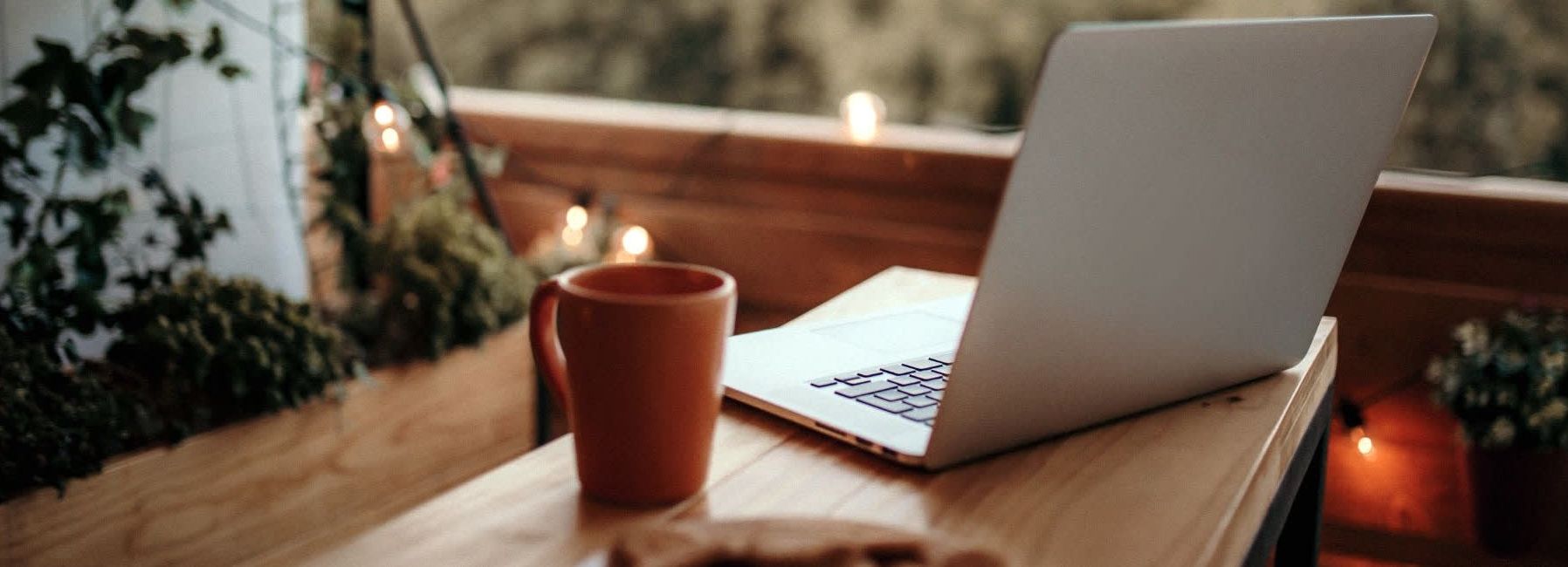 Laptop and coffee mug on a wooden table on a cozy balcony with string lights and plants.